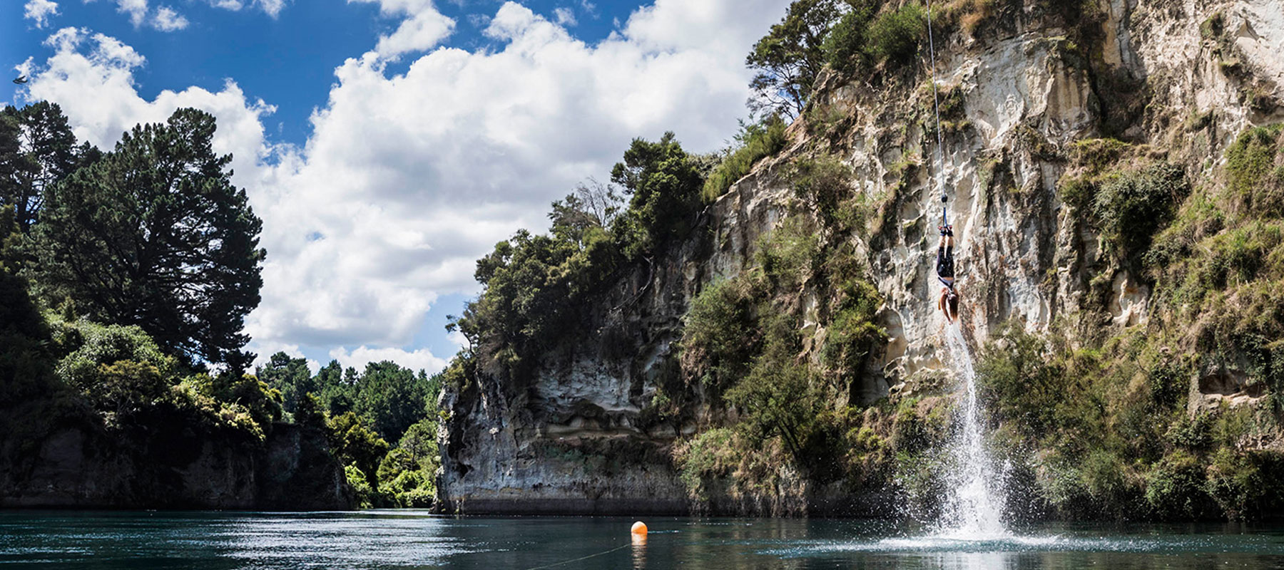 The image shows a person mid-air as they jump off a tall rocky cliff into a calm body of water below. The cliff is rugged, with greenery and trees growing along its edges, and the water splashes beneath the jumper. The surrounding area features more cliff