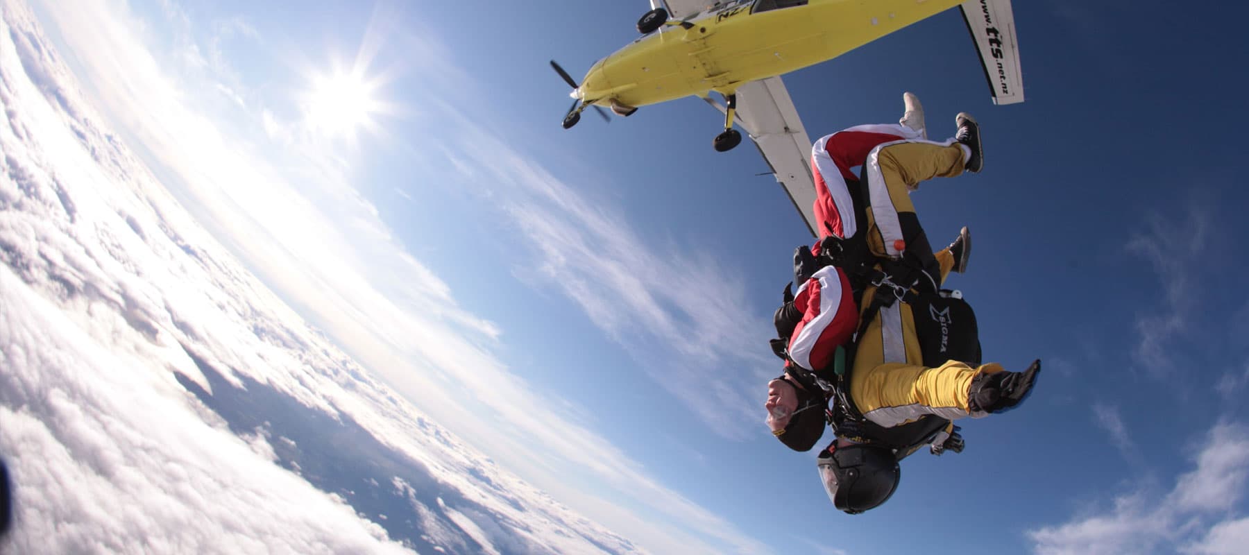 The image captures a thrilling moment of a tandem skydive, with two skydivers in freefall after jumping from a yellow airplane high above the clouds. The skydivers are dressed in jumpsuits, helmets, and parachute gear, with the instructor positioned behin