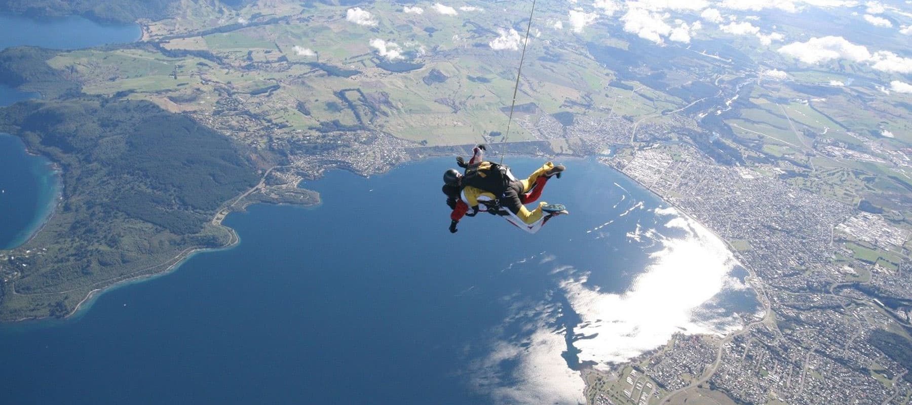  The image shows two tandem skydivers descending above a vast landscape with a large lake and surrounding greenery. The skydivers are suspended mid-air, with the town, water, and rolling hills far below. The sun reflects off the lake’s surface, creating a