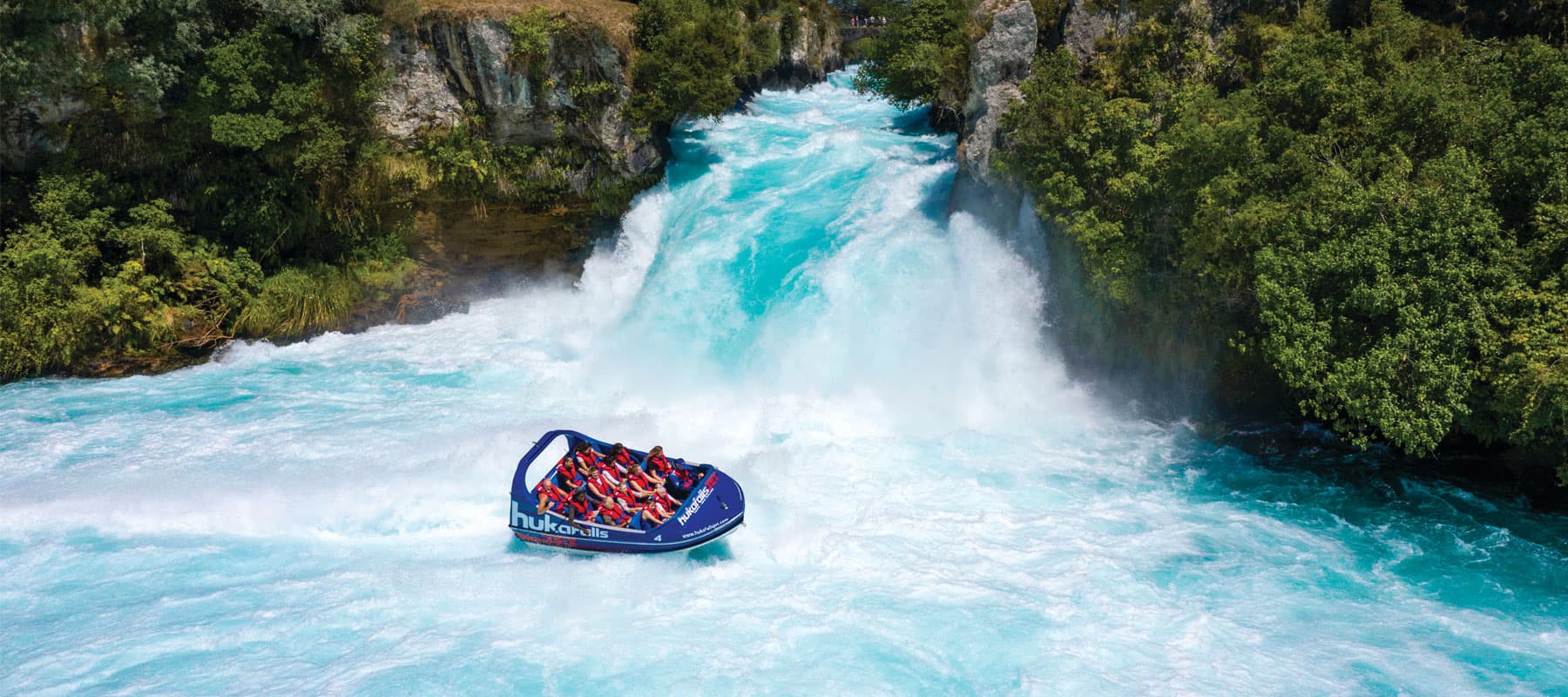 The image shows a blue jet boat labeled "hukafalls" speeding across vibrant blue rapids with white frothy water. The boat, filled with passengers wearing life jackets, navigates near a large waterfall surrounded by rocky cliffs and lush green vegetation. 