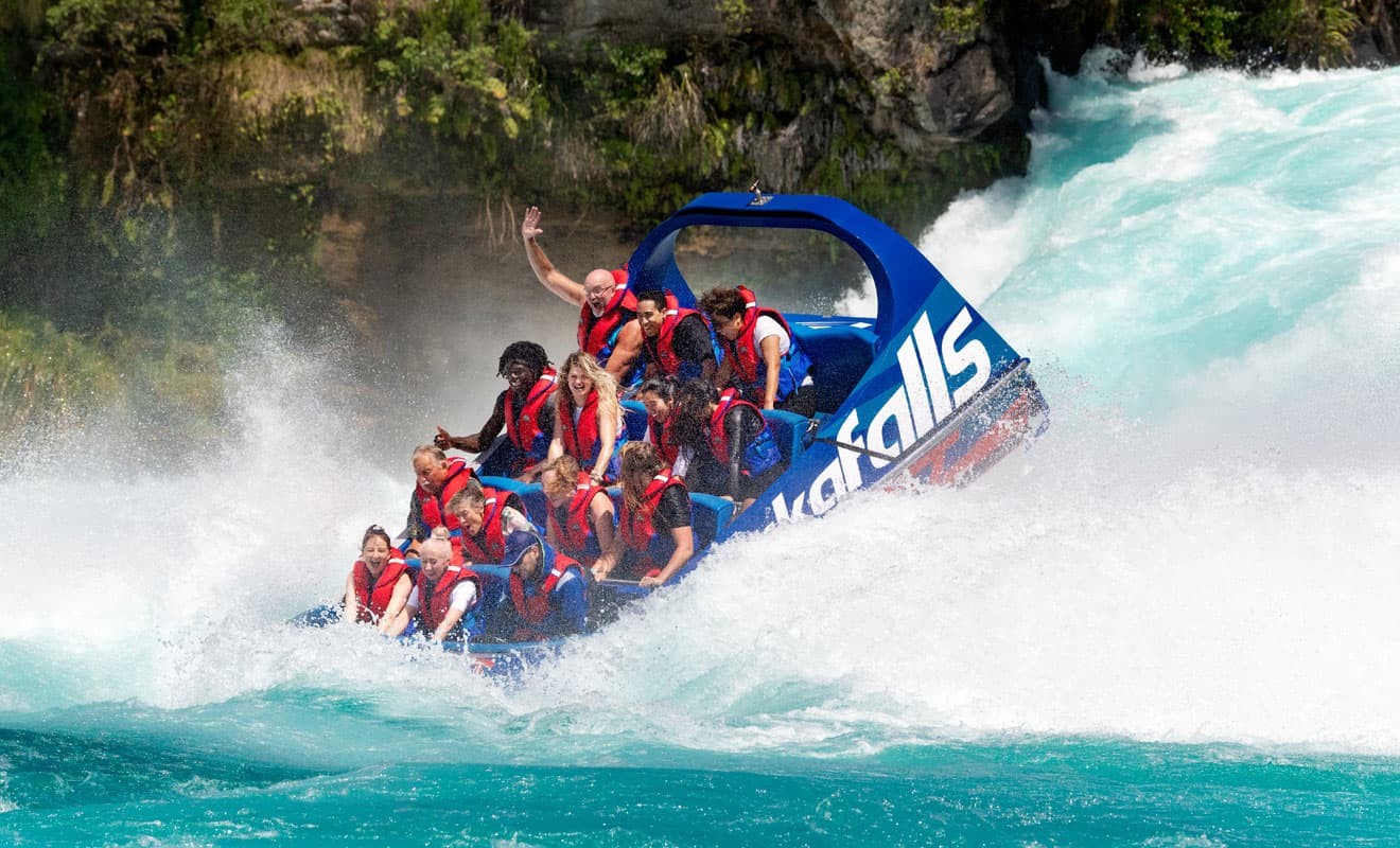 The image shows a blue jet boat labeled "hukafalls" navigating through turbulent whitewater rapids. The passengers, all wearing life jackets, appear to be experiencing the thrill of the ride, with one person raising their hand in excitement. The boat is