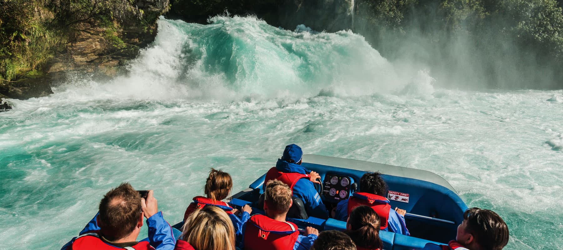 The image shows the back view of a group of passengers wearing red life jackets on a jet boat, approaching a large, powerful waterfall. The passengers are seated, with one person holding up a phone to capture the moment. The water is churning and turbulen