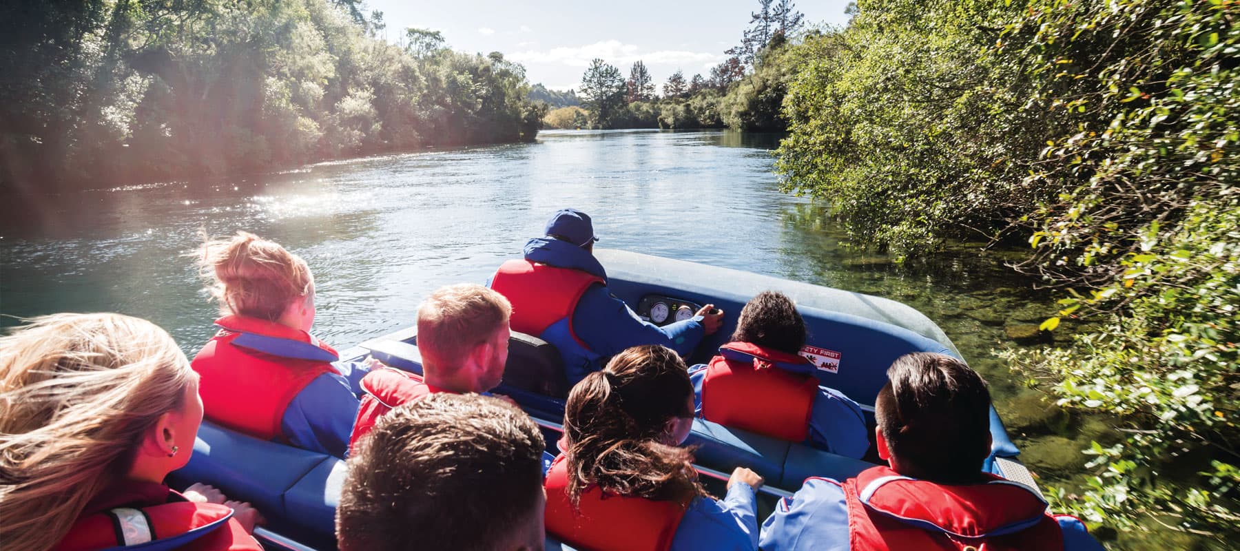  The image shows a group of passengers wearing red life jackets seated on a jet boat, cruising calmly down a serene river. The view is from behind the passengers, with the boat captain steering them through the water. The river is lined with lush green tr