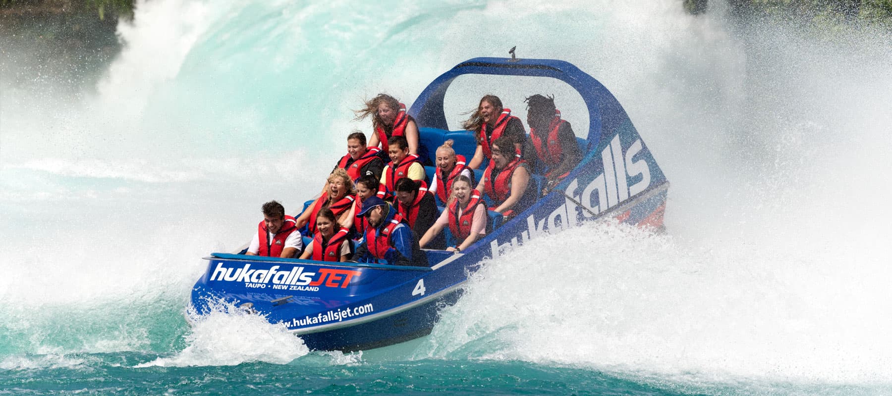 The image shows a blue jet boat labeled "hukafalls JET" filled with excited passengers wearing red life jackets as they navigate through whitewater rapids. The passengers, gripping the boat tightly, appear to be thrilled by the ride, with expressions of j