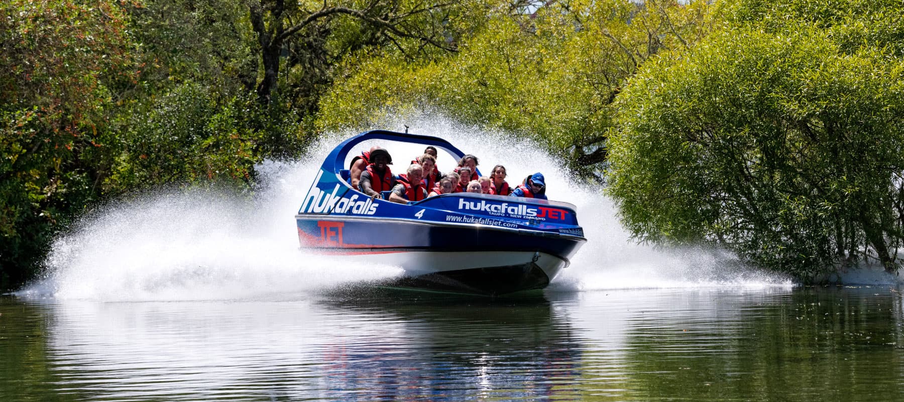 The image shows a blue jet boat labeled "hukafalls JET" speeding through calm water, creating large splashes on both sides as it cuts through the surface. The boat is filled with passengers wearing red life jackets, holding on as they experience the thril