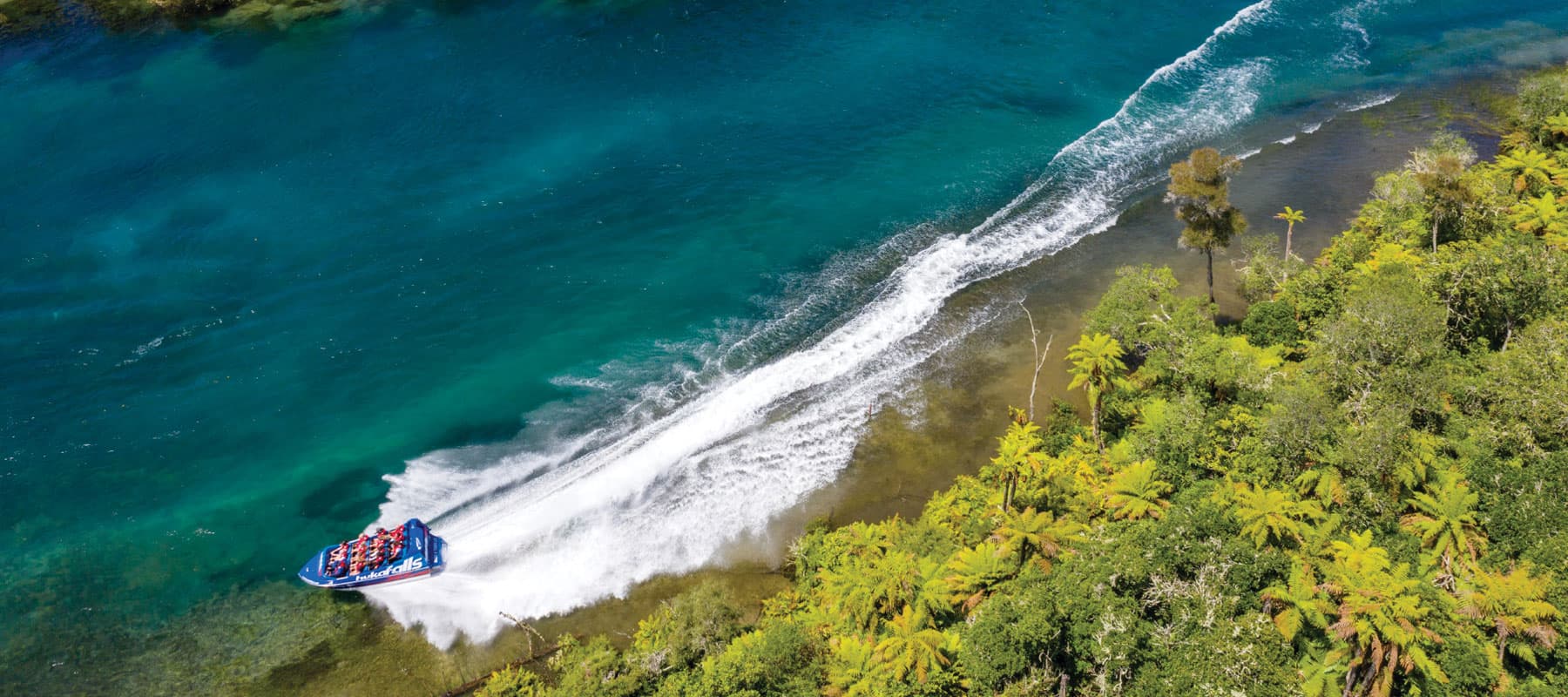 The image shows an aerial view of a blue jet boat labeled "hukafalls" speeding along a clear, turquoise river near the shoreline. The boat leaves a large white wake behind as it zips through the water. On the right, lush green tropical trees and plants li