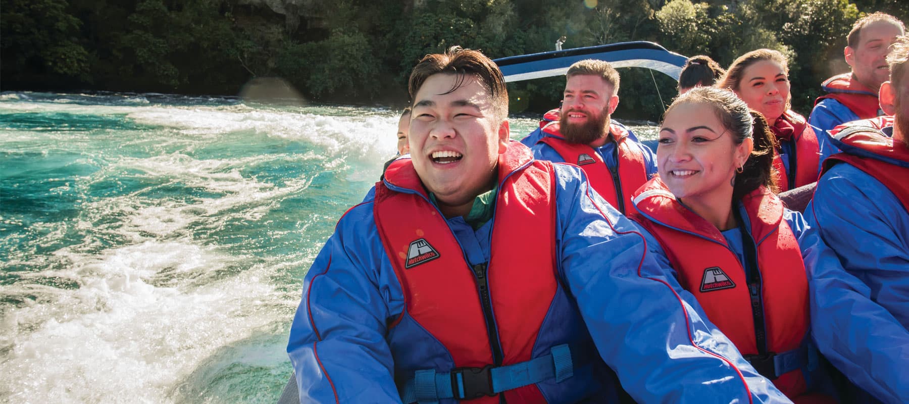 The image shows a group of passengers wearing red life jackets and blue jackets, enjoying a thrilling ride on a jet boat. The focus is on a smiling man at the front, who is clearly exhilarated by the experience, while the passengers behind him also appear