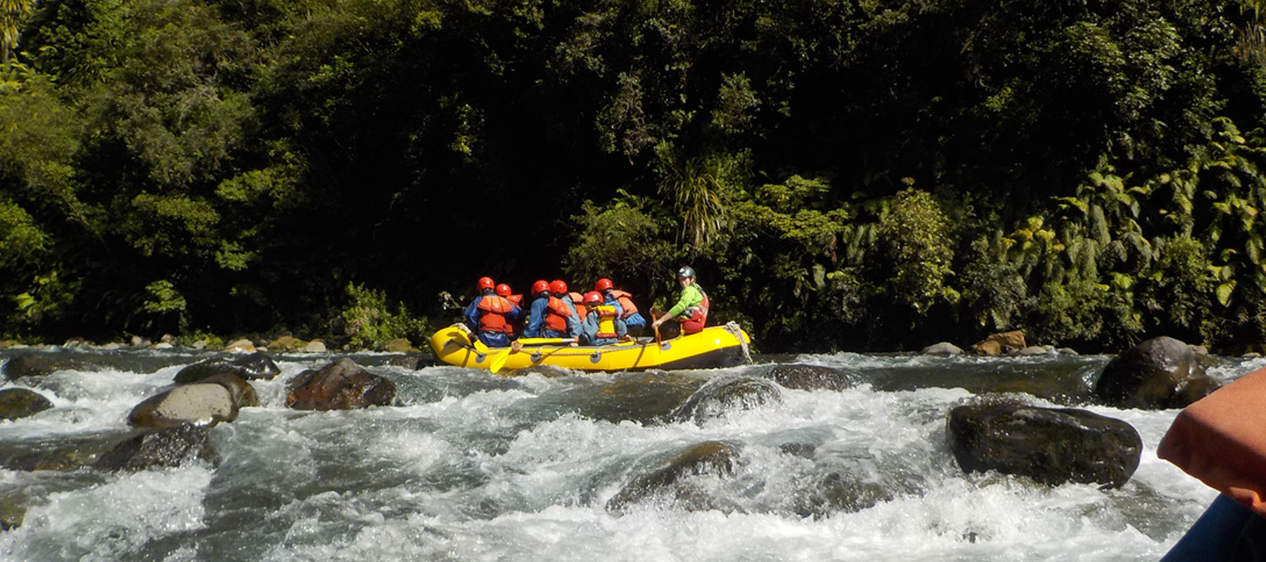 The image shows a group of people rafting down a river in a yellow inflatable raft. The passengers, wearing orange life jackets and helmets, are navigating through rocky rapids, with whitewater surrounding the raft. A guide at the back steers the raft wh