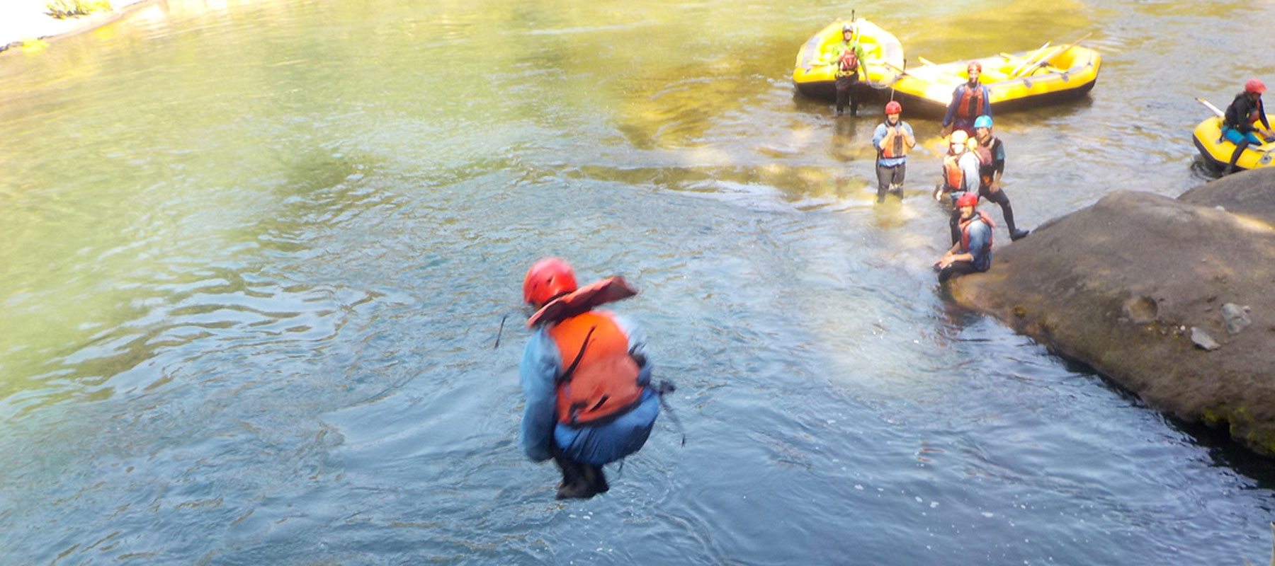 The image shows a person wearing an orange life jacket and red helmet mid-jump into a calm river, creating a splash. In the background, a group of people in similar gear stand near the water's edge, with yellow inflatable rafts visible on the riverbank. 