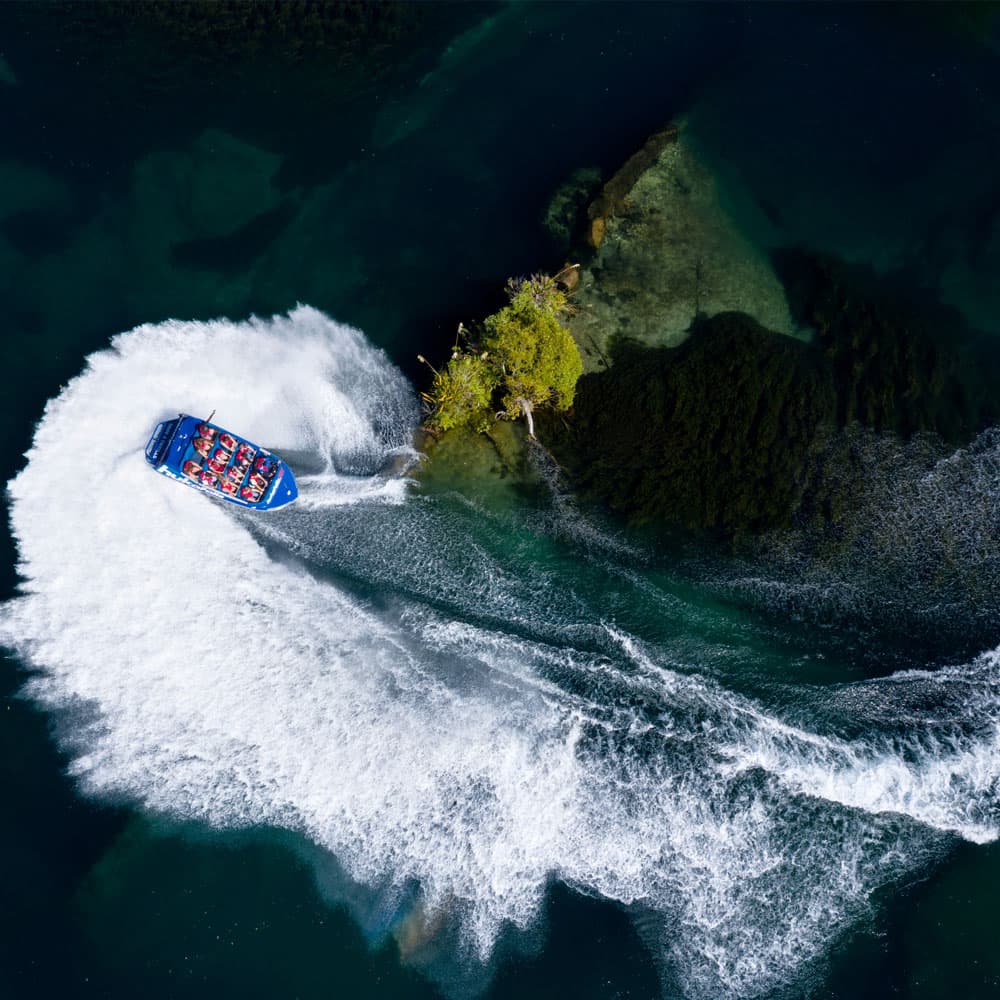  The image shows an aerial view of a blue jet boat labeled "hukafalls JET" making a sharp turn near a small, rocky outcrop surrounded by dark blue-green water. The boat creates a large, white spray of water as it maneuvers through the river, leaving a swi