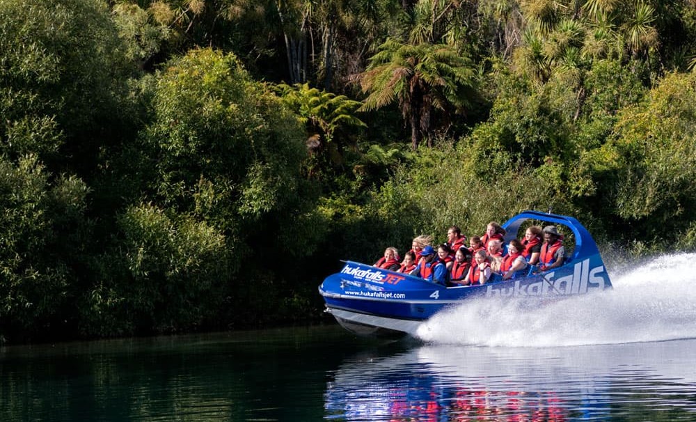 The image shows a blue jet boat labeled "hukafalls JET" speeding along a calm river, with lush greenery and trees lining the riverbank. The boat is filled with passengers wearing red life jackets, holding on as the boat creates a large spray of water behi