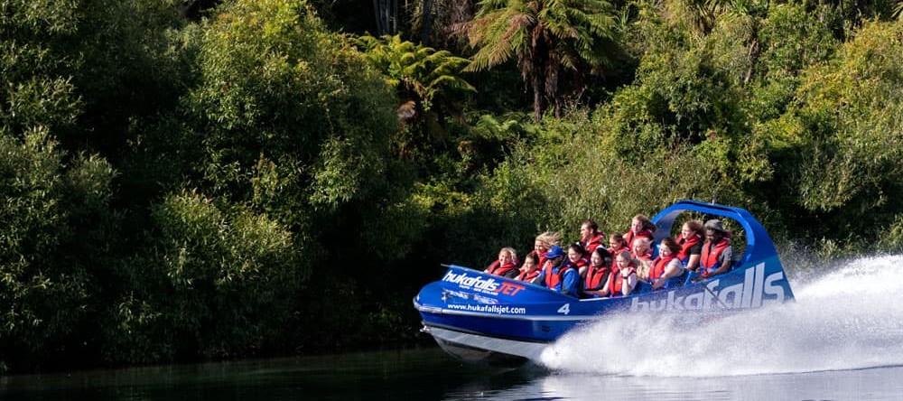 The image shows a blue jet boat labeled "hukafalls JET" speeding along a calm river, with lush greenery and trees lining the riverbank. The boat is filled with passengers wearing red life jackets, holding on as the boat creates a large spray of water behi